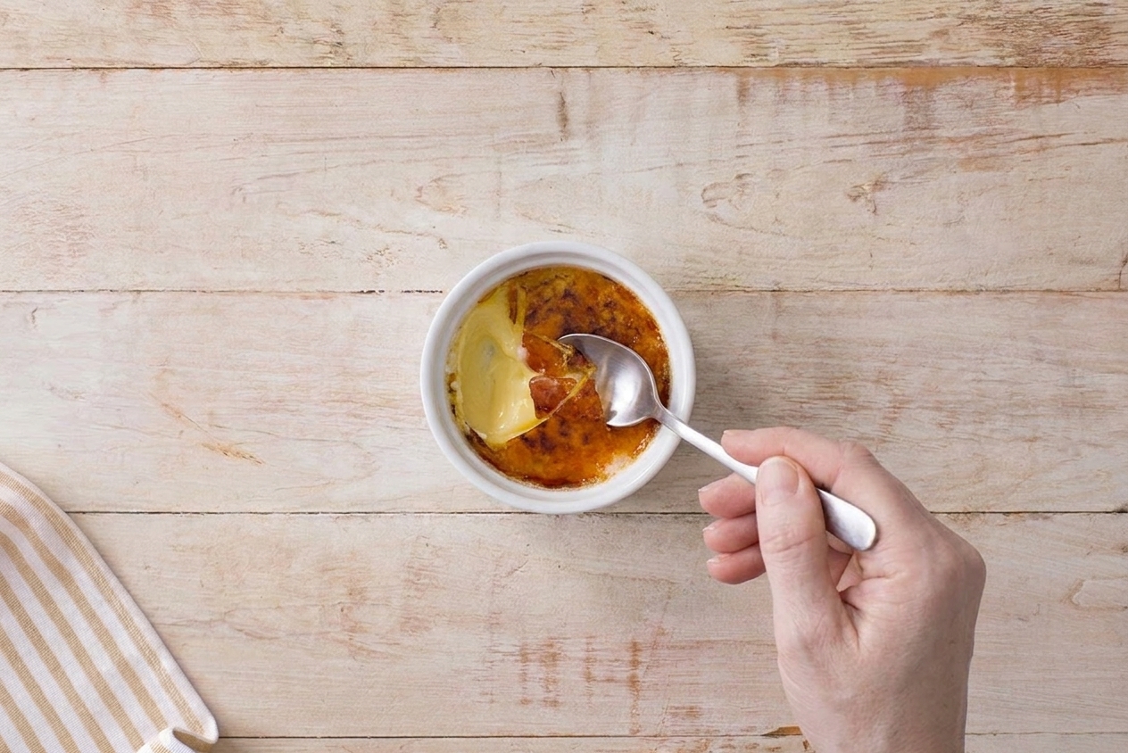 A spoon breaking through the caramelized sugar crust of a crème brûlée, showing creamy custard underneath