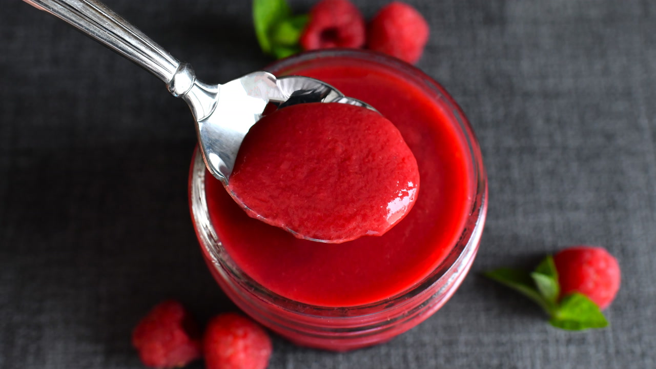 A spoon drizzling glossy deep-red raspberry sauce over a slice of cheesecake on a white plate on a bright kitchen table