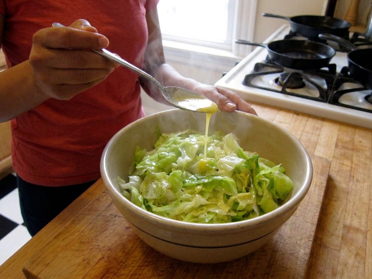 A spoon drizzling melted butter over hot boiled cabbage in a serving bowl on a kitchen counter
