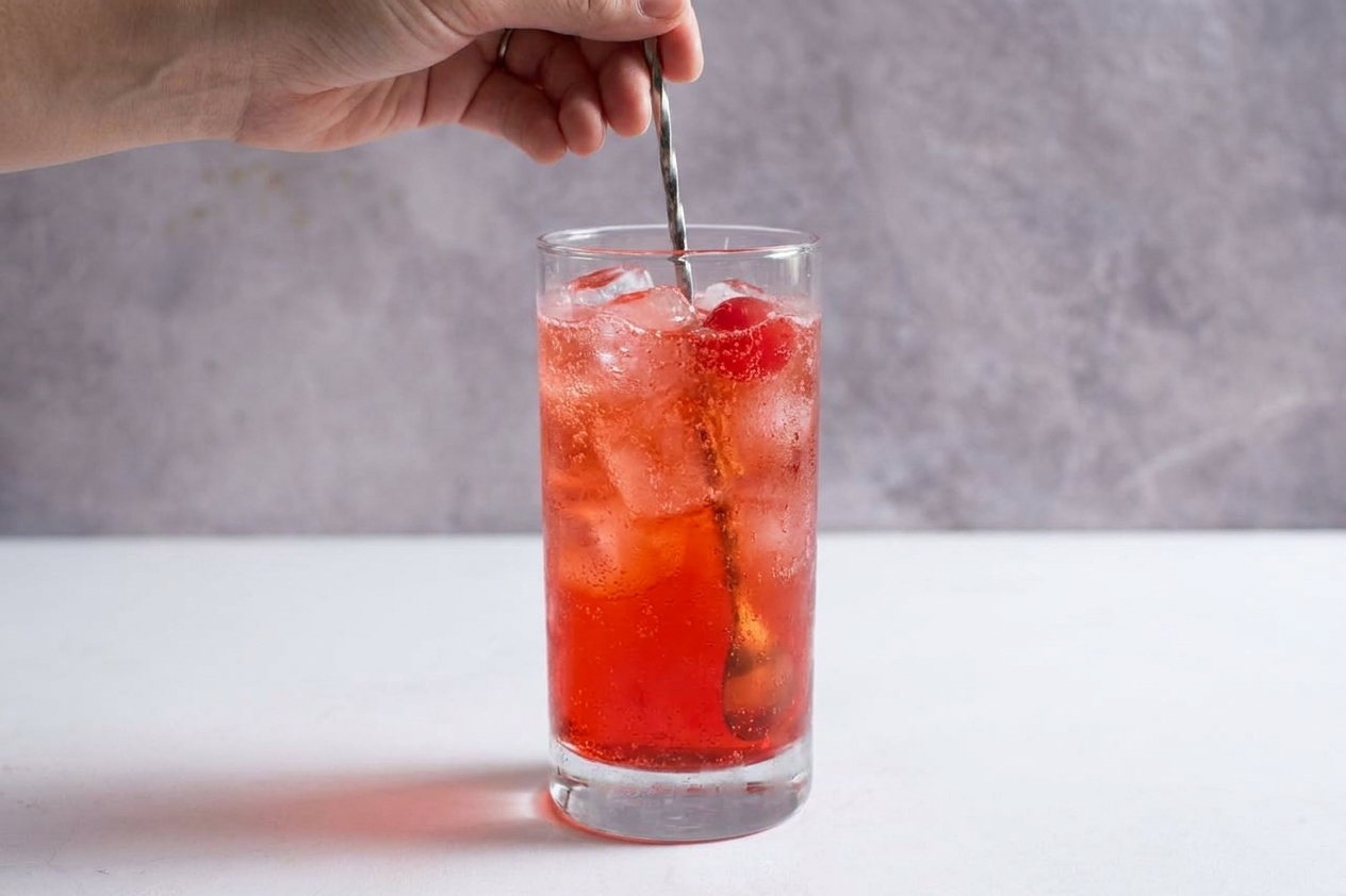 A spoon gently stirring a fizzy Shirley Temple with ice in a tall glass on a bright kitchen counter