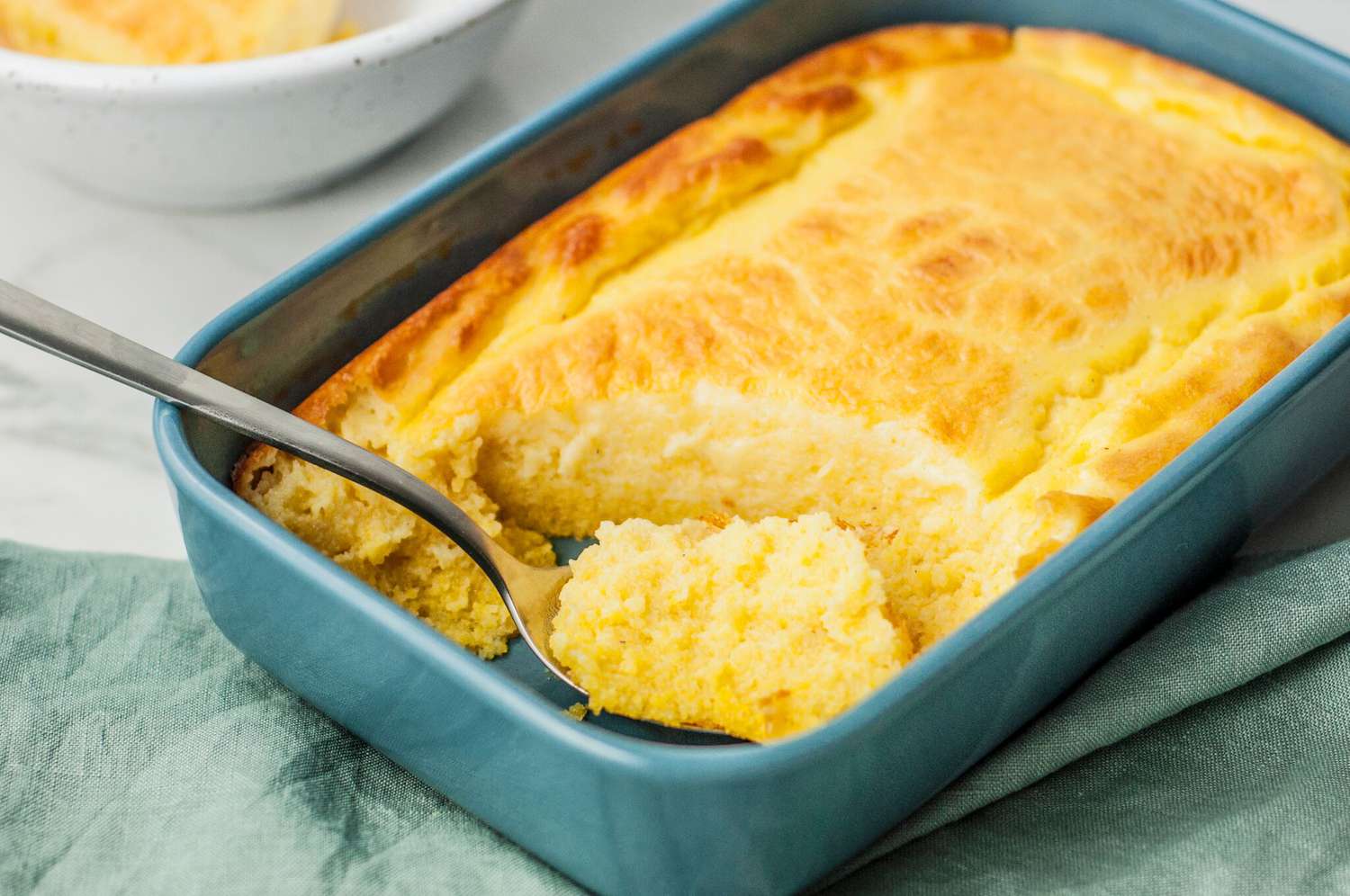 A spoon scooping a warm serving of cornbread pudding from a baking dish on a bright, neutral kitchen counter