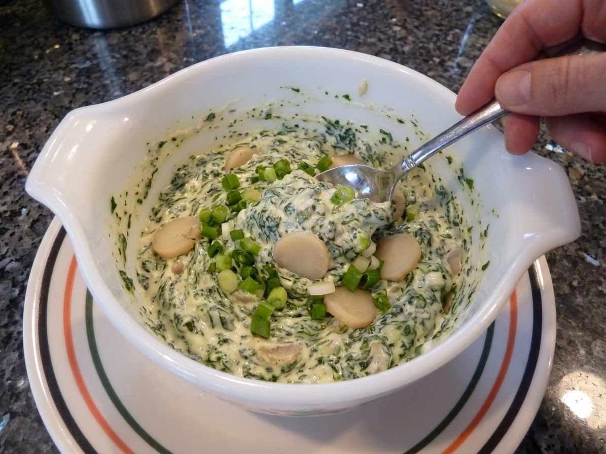 A spoon stirring creamy spinach dip in a white mixing bowl with green onions and water chestnuts visible