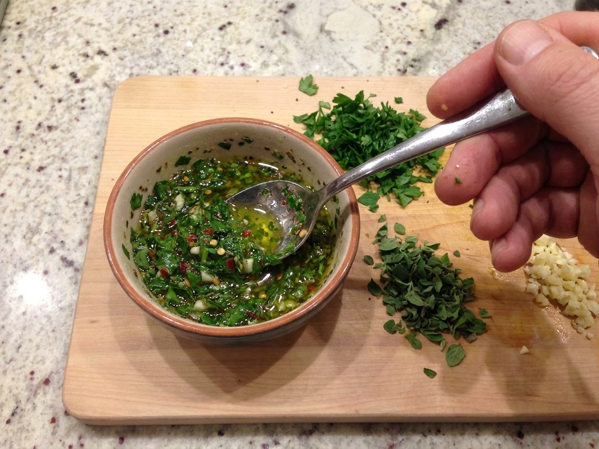 A spoon stirring fresh green chimichurri in a small bowl on a kitchen counter, with chopped herbs nearby