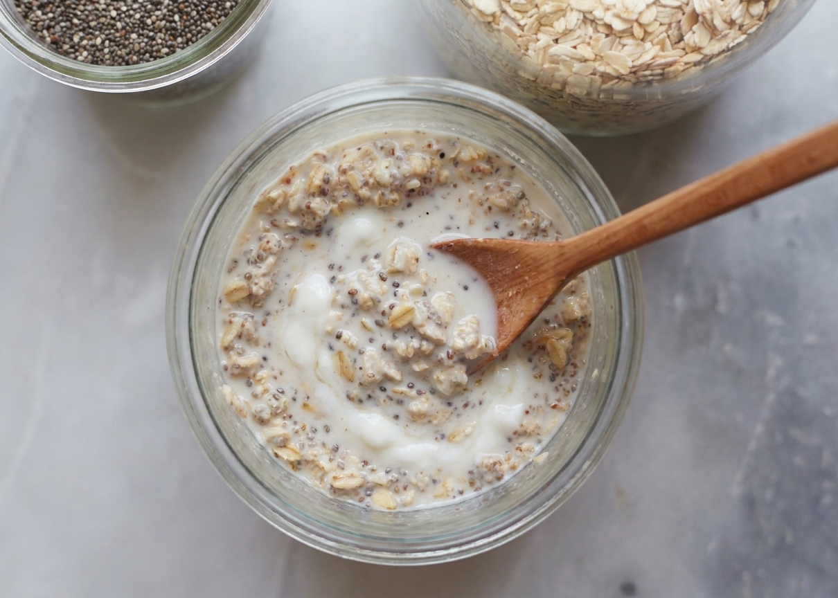 A spoon stirring oats and chia seeds with milk and yogurt in a glass jar on a kitchen counter
