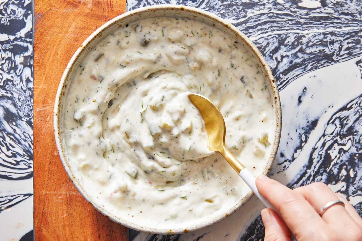 A spoon stirring tartar sauce in a small white bowl, showing pickle and herb flecks throughout