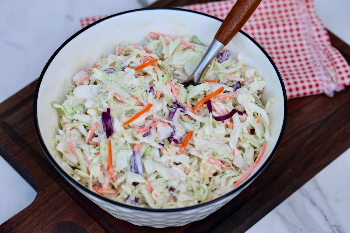A spoon tossing creamy coleslaw in a large mixing bowl on a kitchen counter, close-up real food photography style