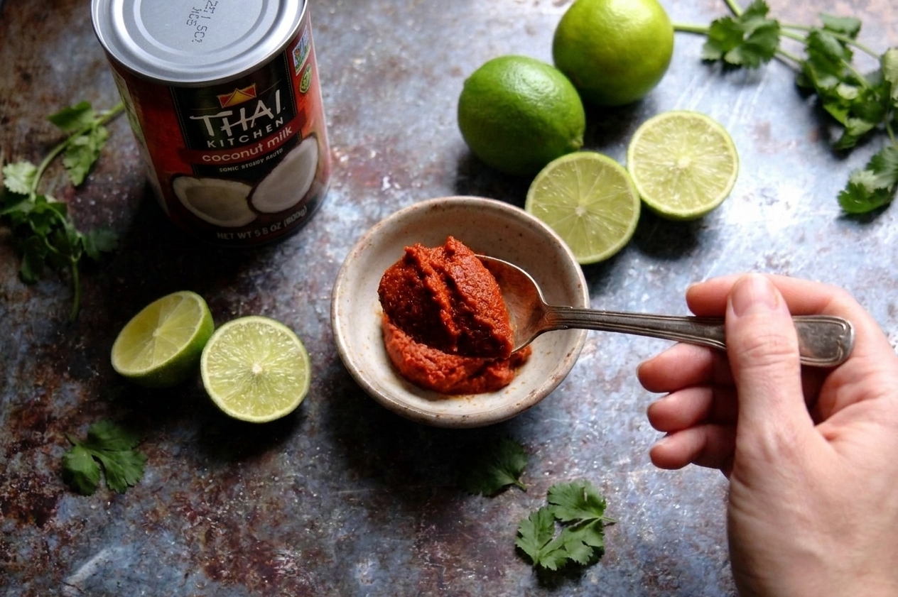 A spoonful of Thai red curry paste held above a small bowl on a bright kitchen counter, with coconut milk and lime nearby