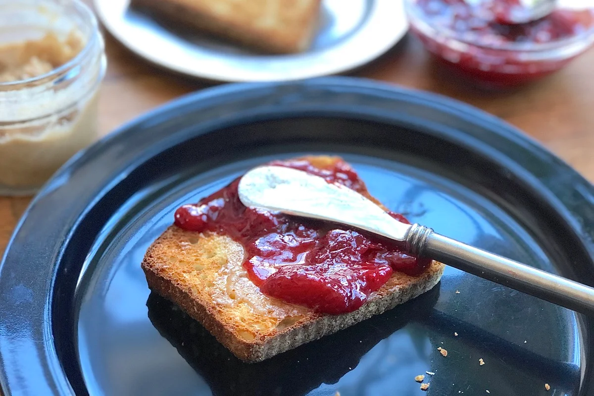 A spoonful of strawberry freezer jam being spread onto toasted bread on a simple ceramic plate at a kitchen table in soft daylight