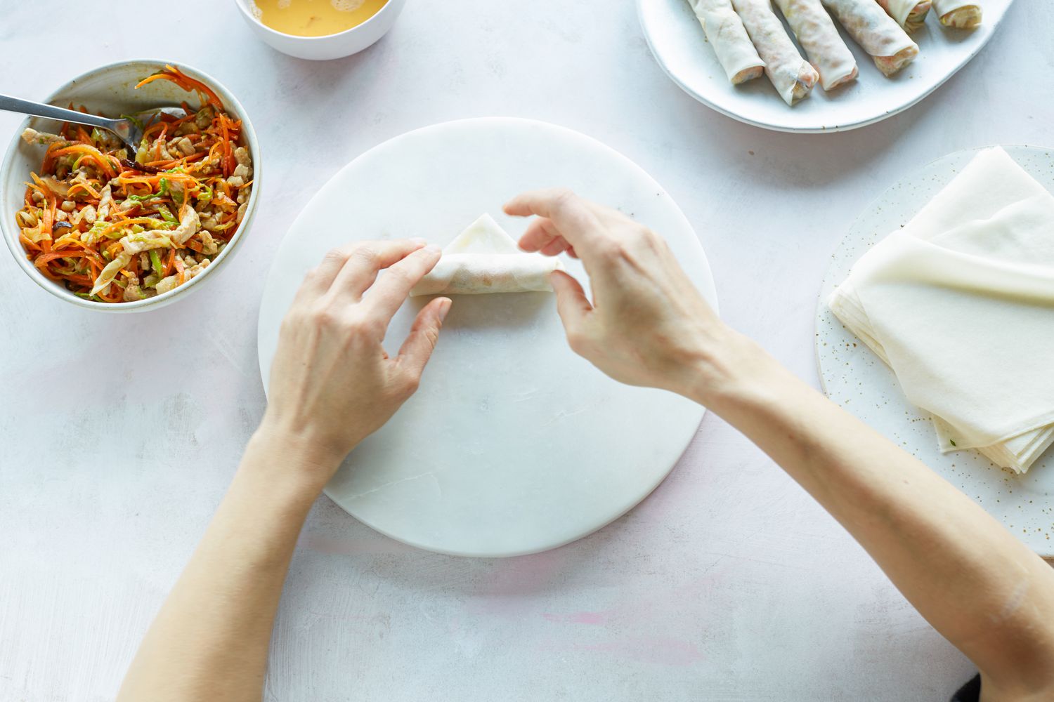 A spring roll rolling station with wrappers under a towel, a bowl of filling, a small bowl of flour slurry, and a sheet pan ready for rolled spring rolls on a kitchen counter
