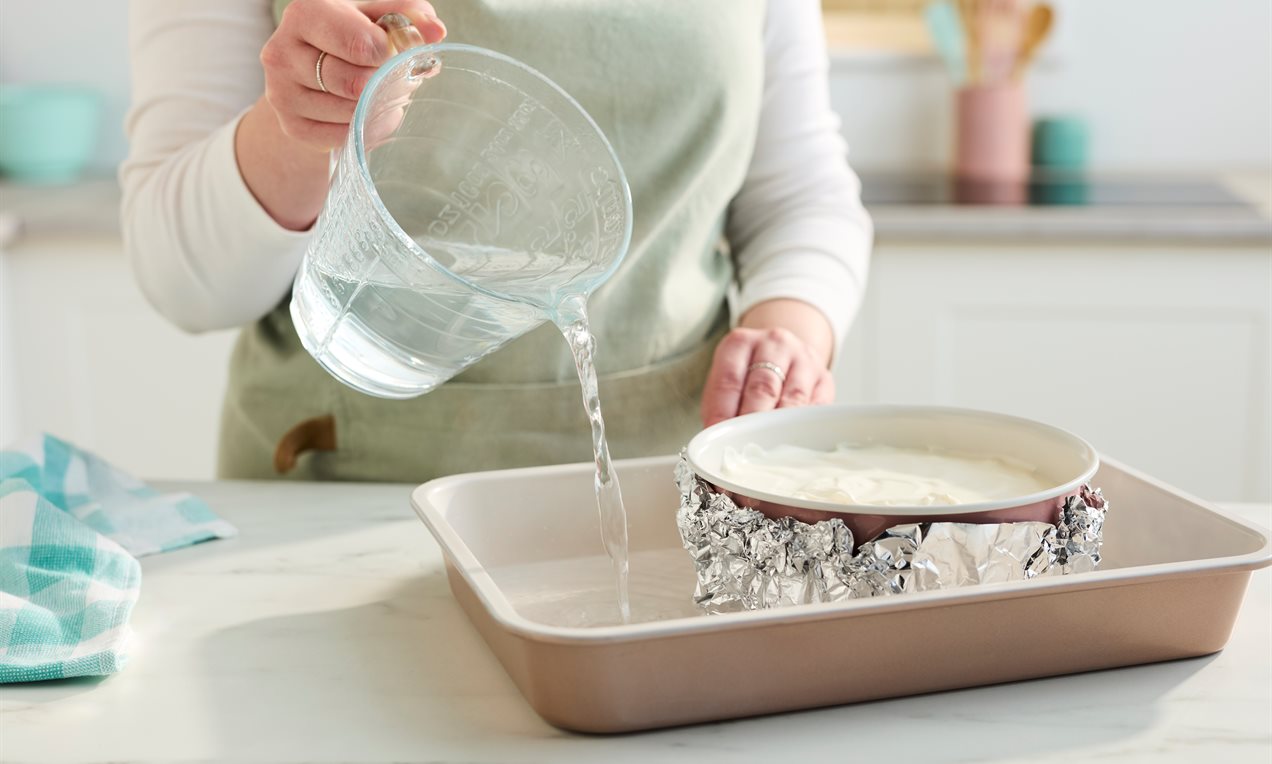 A springform pan wrapped in foil sitting in a roasting pan filled with hot water inside an oven, mid-bake