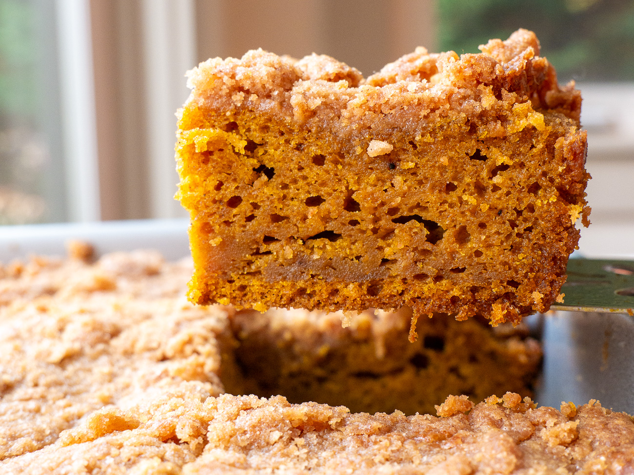 A square baking pan with pumpkin cake batter being spread with a spatula, with streusel crumbs nearby on the counter