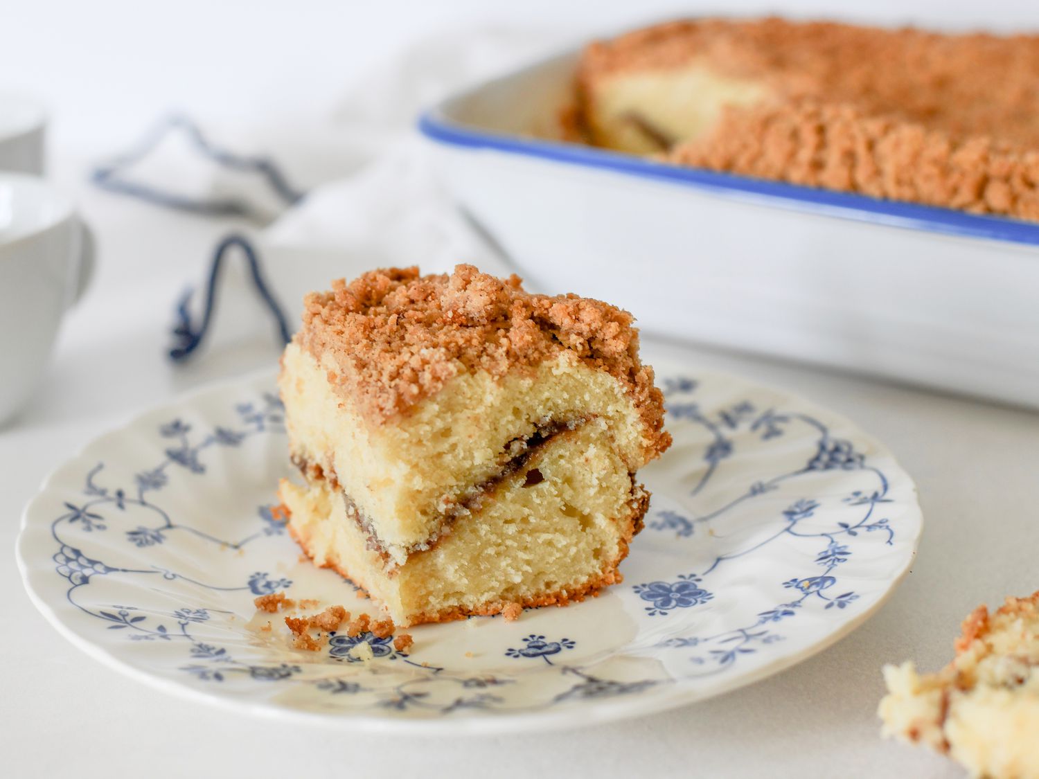 A square cinnamon coffee cake in a baking pan with a thick crumb topping and a few slices cut out, on a warm wooden kitchen counter in natural morning light