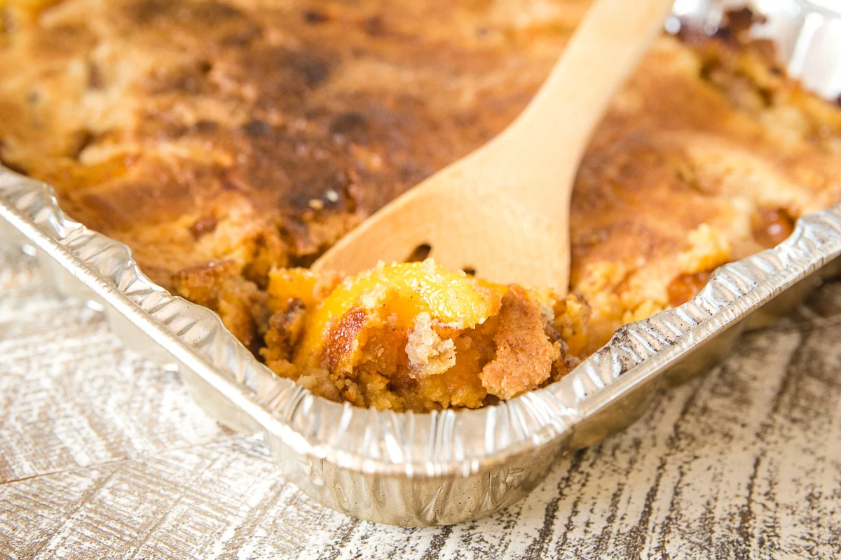 A square of peach cobbler in a clear airtight container with a spoon beside it on a kitchen counter