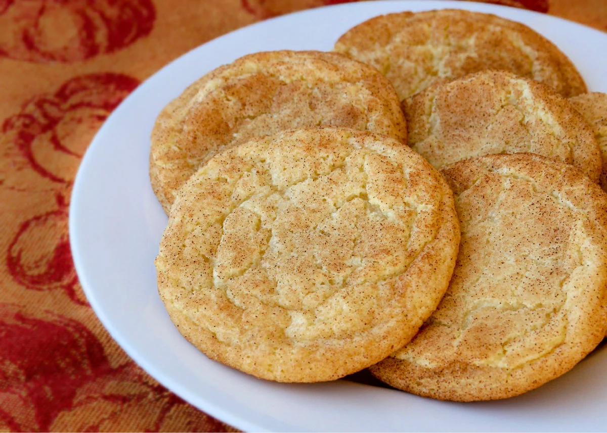 A stack of cinnamon-sugar snickerdoodle cookies on a ceramic plate on a warm-toned kitchen table with soft natural window light