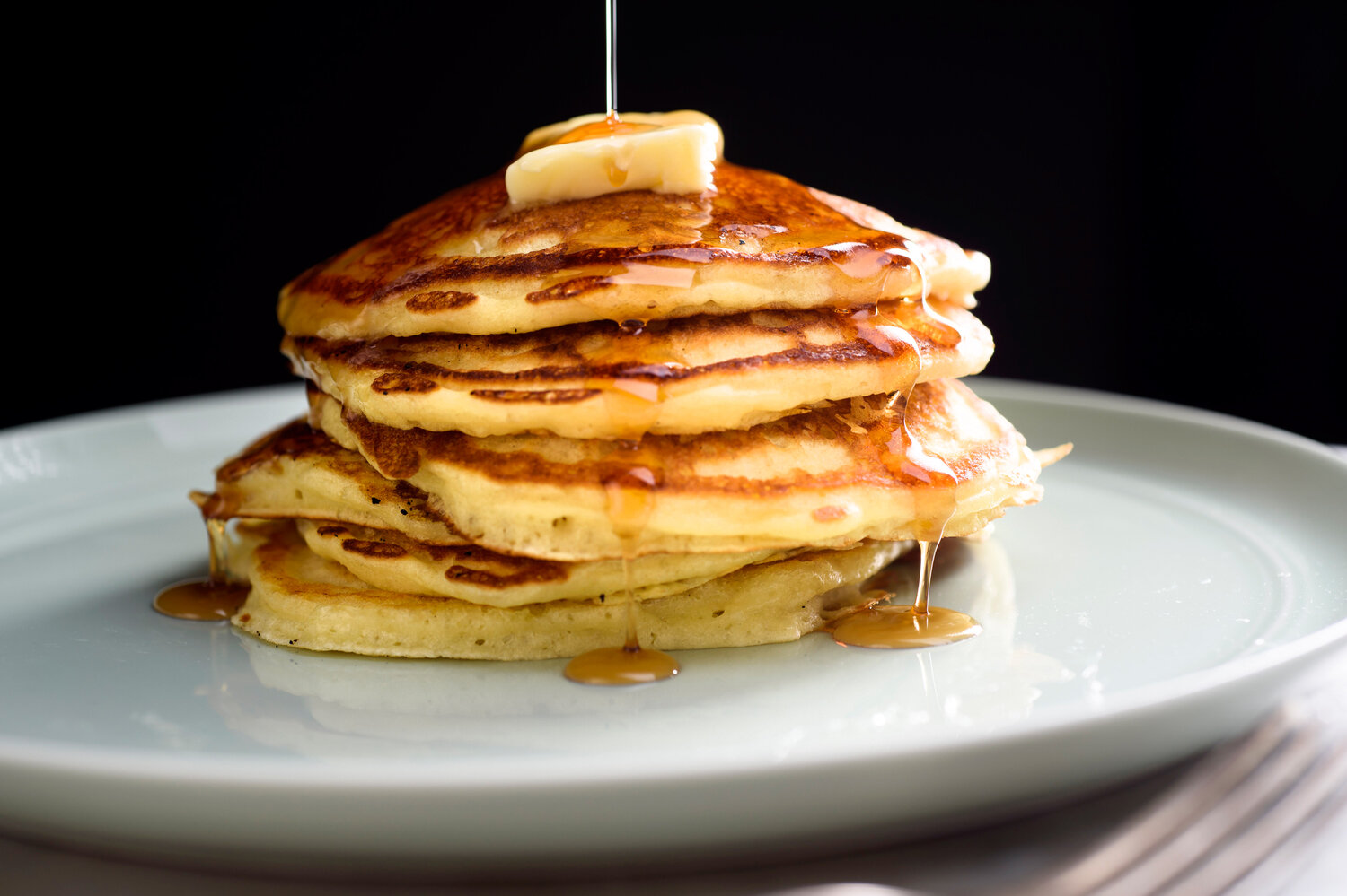 A stack of fluffy golden pancakes on a white plate with butter melting on top and maple syrup nearby on a bright kitchen table