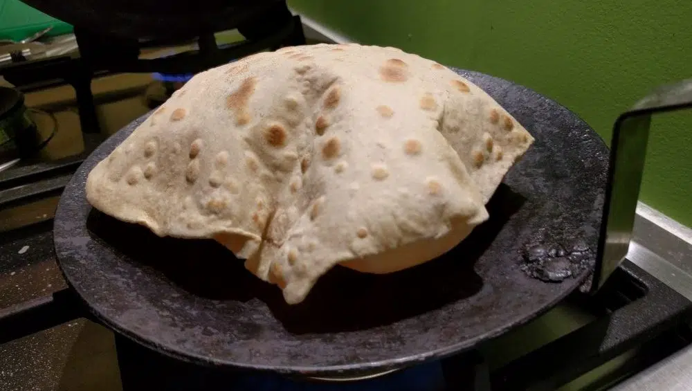 A stack of fresh chapati on a plate beside a hot tawa on a home stove, with one chapati puffed and lightly blistered