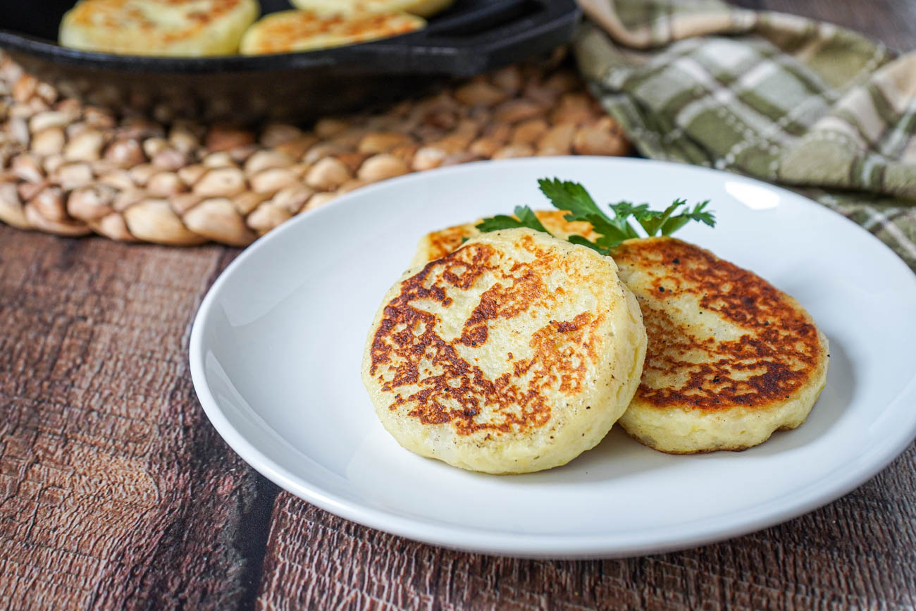 A stack of golden Irish potato cakes on a white plate with a small bowl of sour cream and sliced chives on a warm kitchen table