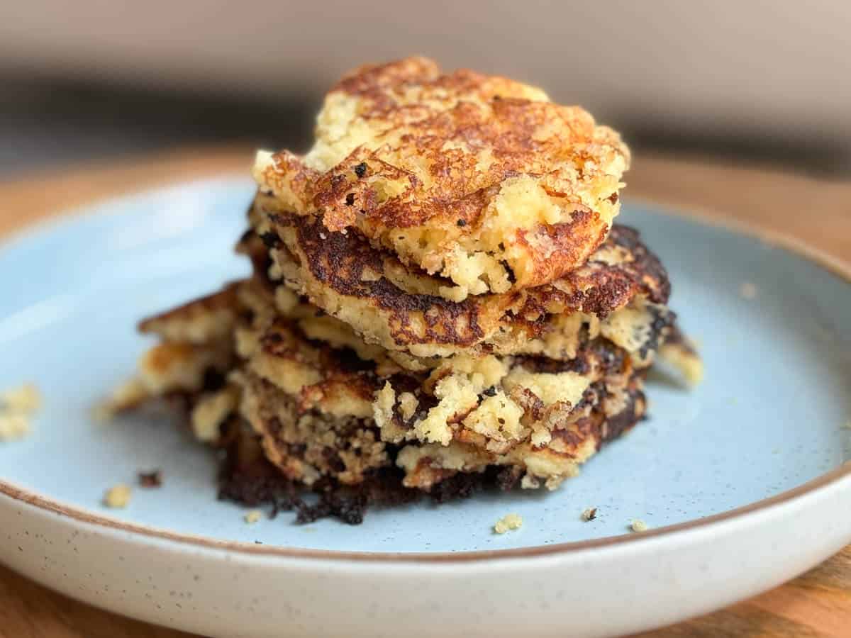 A stack of golden Johnny cakes with crisp browned edges on a plate, with a pat of butter melting on top on a rustic kitchen table