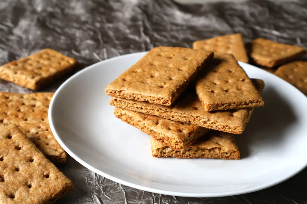 A stack of golden brown homemade graham wafers on a white plate on a sunlit kitchen table, with a few crumbs scattered nearby