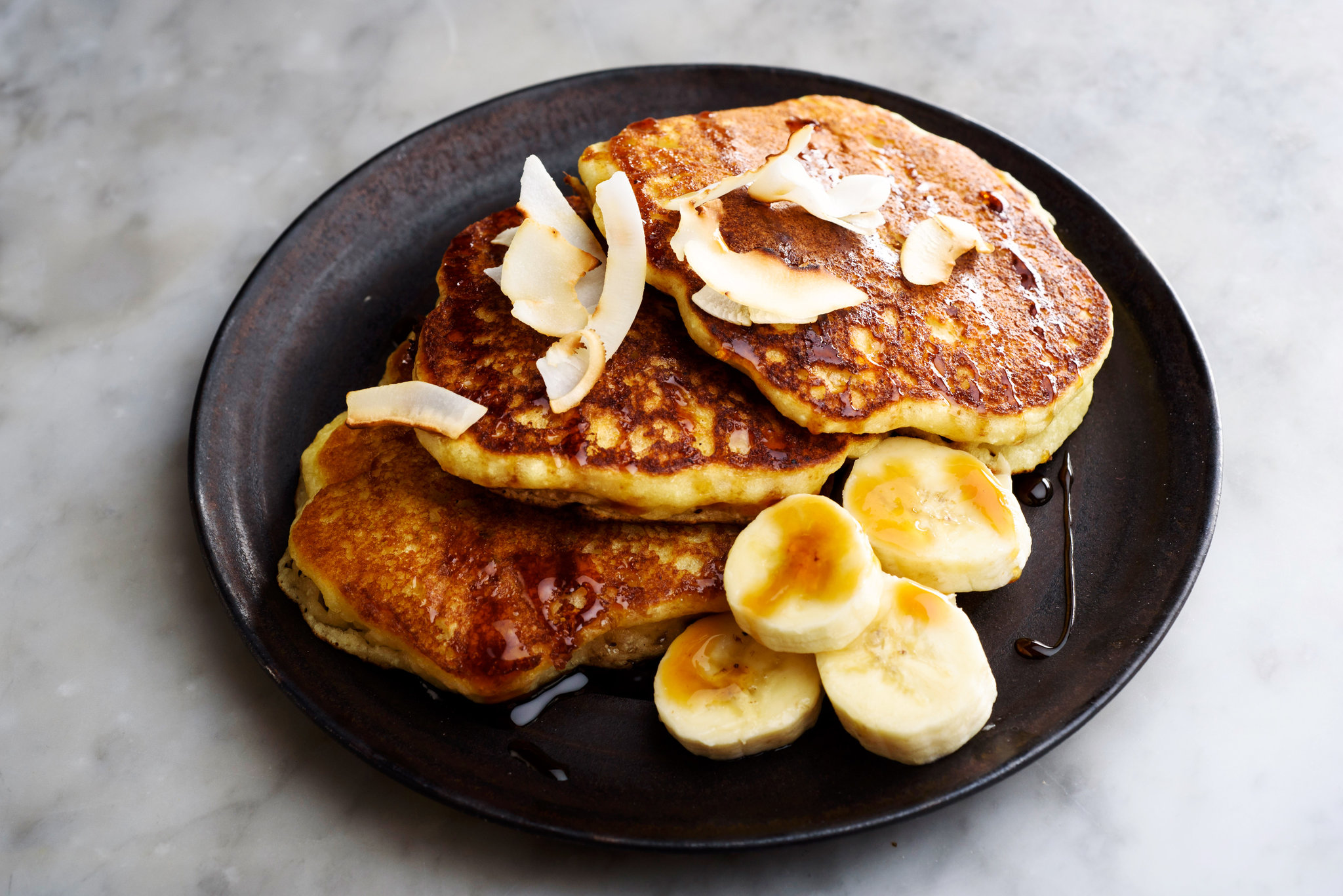 A stack of golden coconut flour pancakes on a white plate with butter melting on top and a drizzle of maple syrup, set on a bright kitchen table