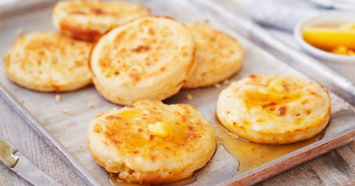 A stack of golden homemade crumpets on a ceramic plate with butter melting into the holes and a small dish of honey on a wooden table in soft morning light
