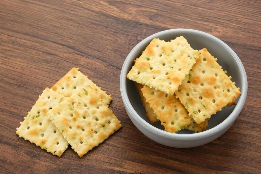 A stack of square hardtack crackers with docked holes on a wooden cutting board on a bright kitchen counter