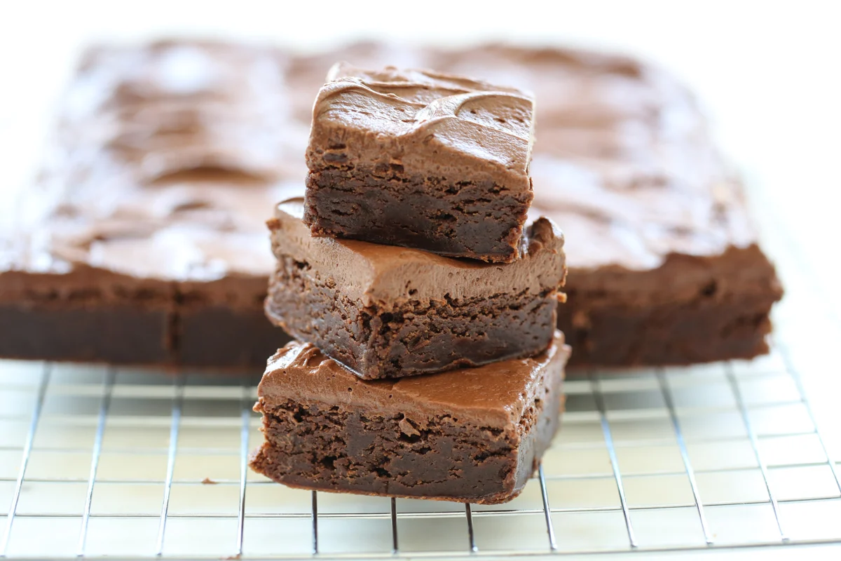 A stack of thick fudgy brownies with a shiny crackly top on a white plate on a warm-toned kitchen table