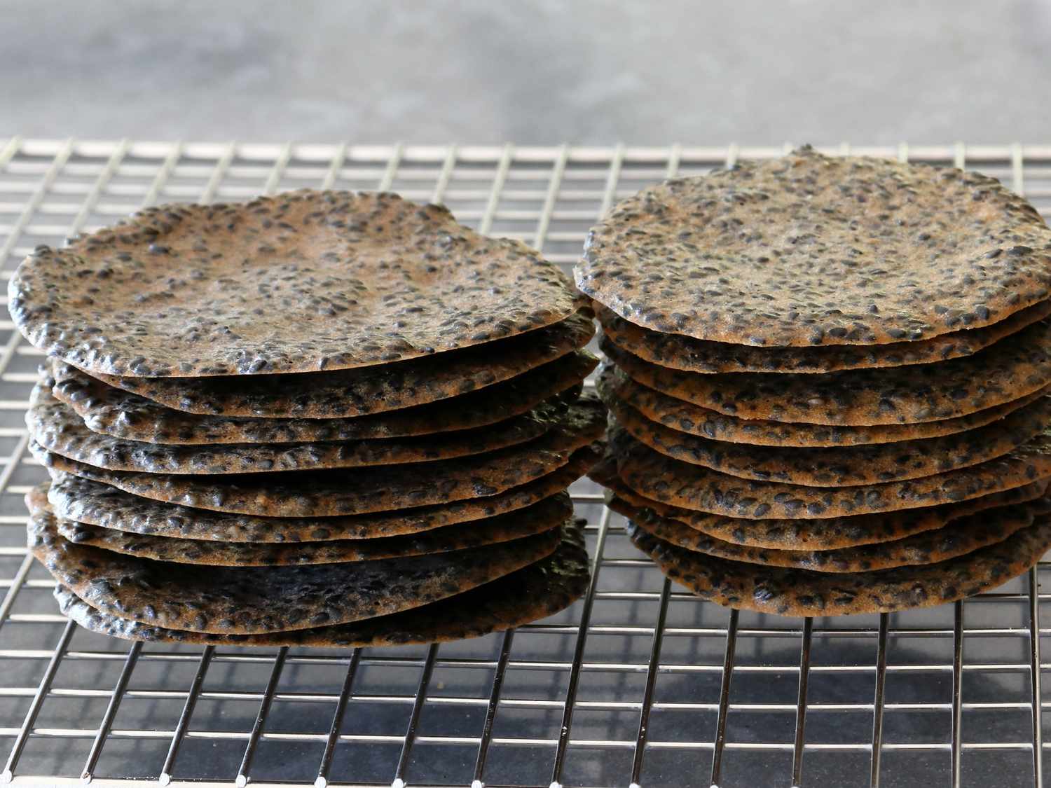 A stack of thin, golden homemade rice crackers sprinkled with sesame seeds on a wooden board with a small bowl of soy sauce nearby, warm kitchen lighting
