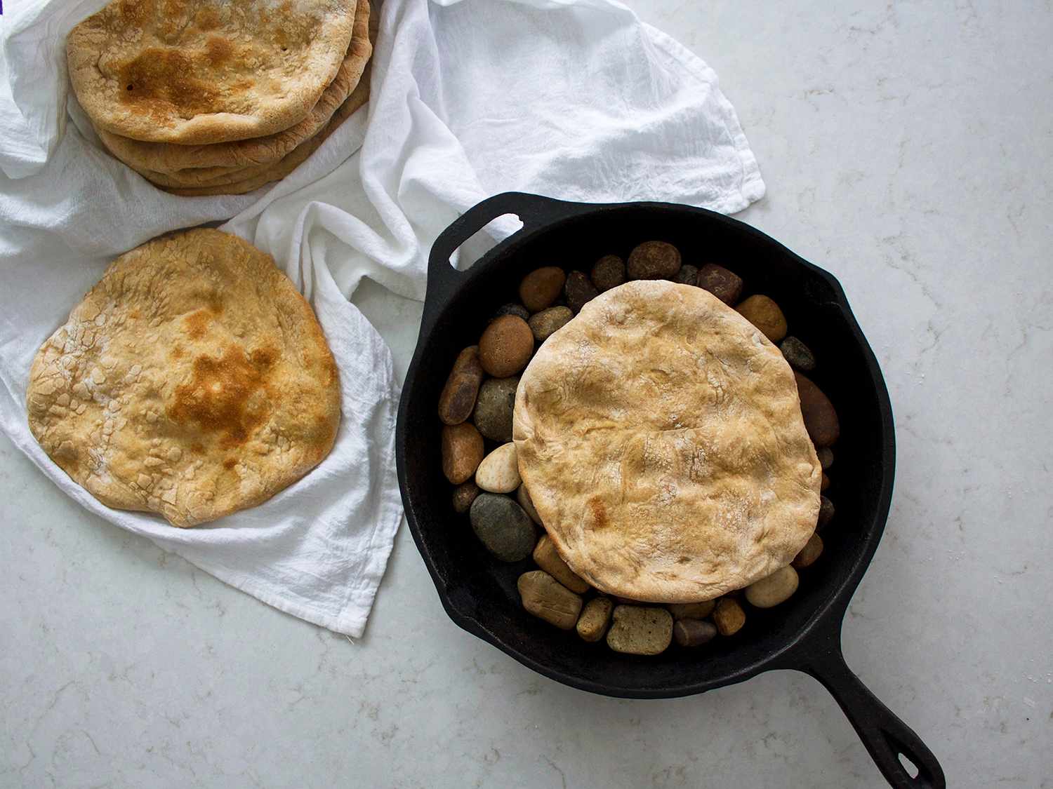 A stack of warm unleavened flatbreads on a wooden cutting board beside a cast iron skillet on a bright kitchen counter