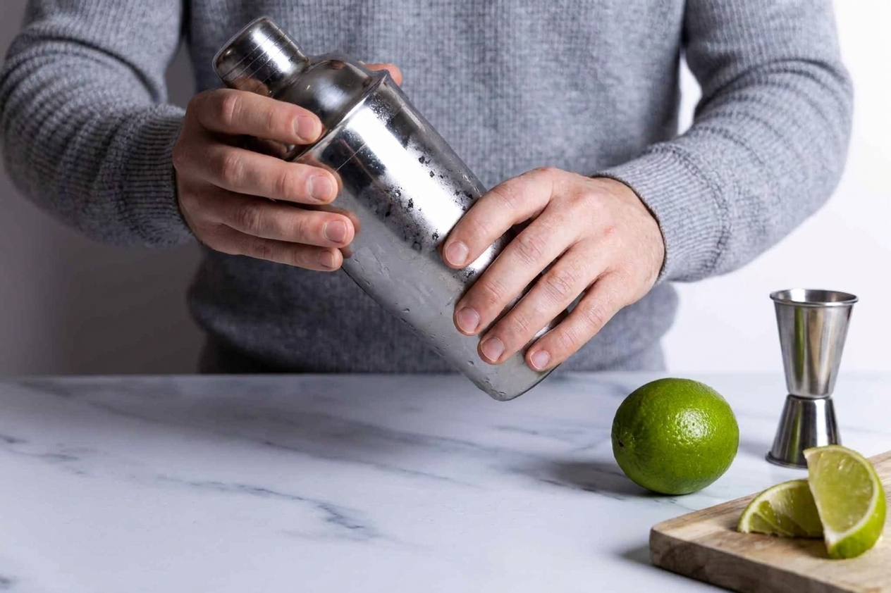 A stainless steel cocktail shaker being shaken over a kitchen counter with a lime and jigger nearby
