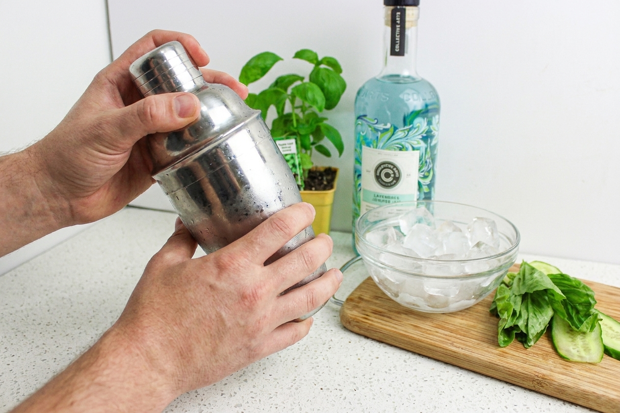 A stainless steel cocktail shaker being shaken over a kitchen counter with a bowl of ice nearby