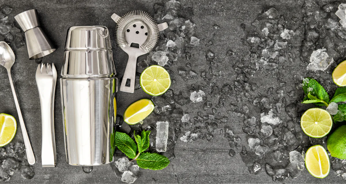 A stainless steel cocktail shaker next to whole lemons and limes on a home kitchen counter