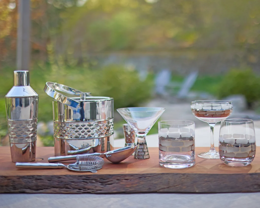 A stainless steel cocktail shaker with a jigger and fine-mesh strainer arranged on a clean kitchen counter under warm indoor lighting