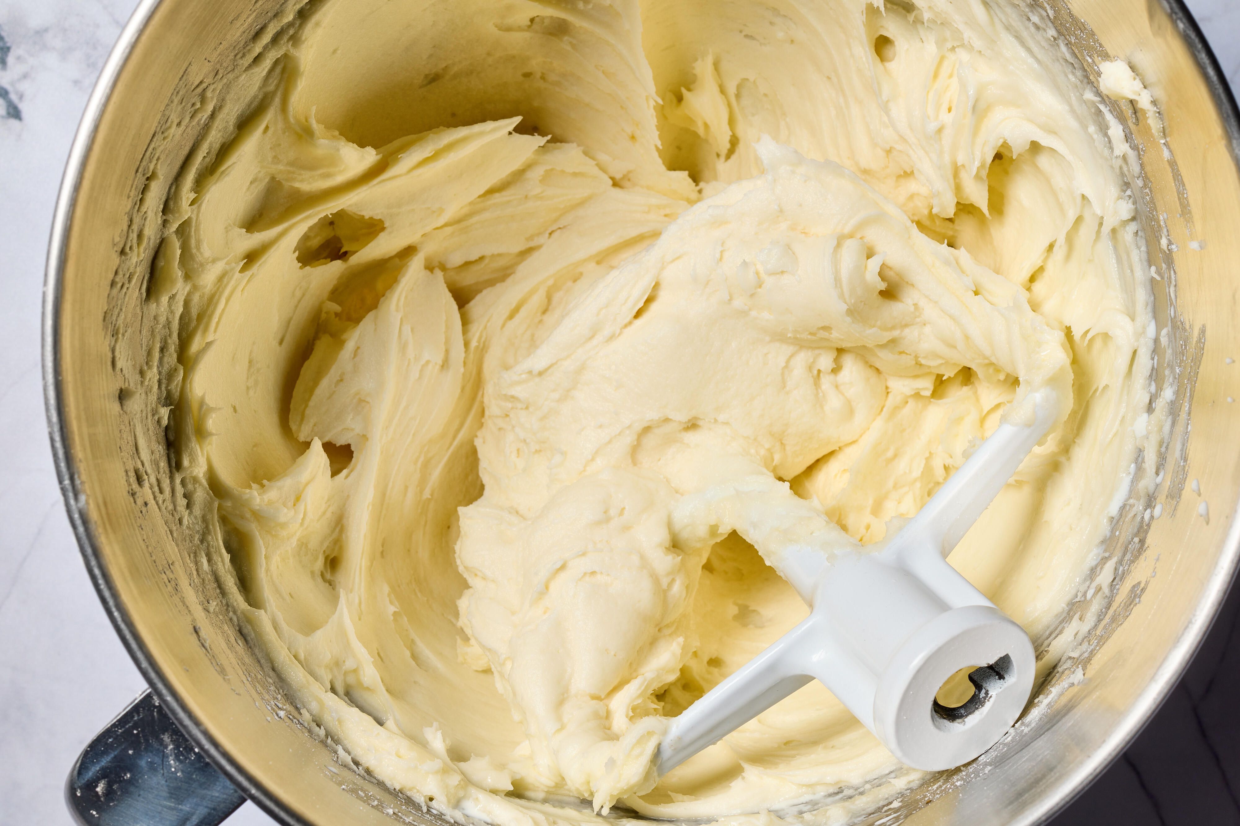 A stainless steel mixing bowl with cream cheese icing being whipped by a hand mixer, with smooth creamy texture forming
