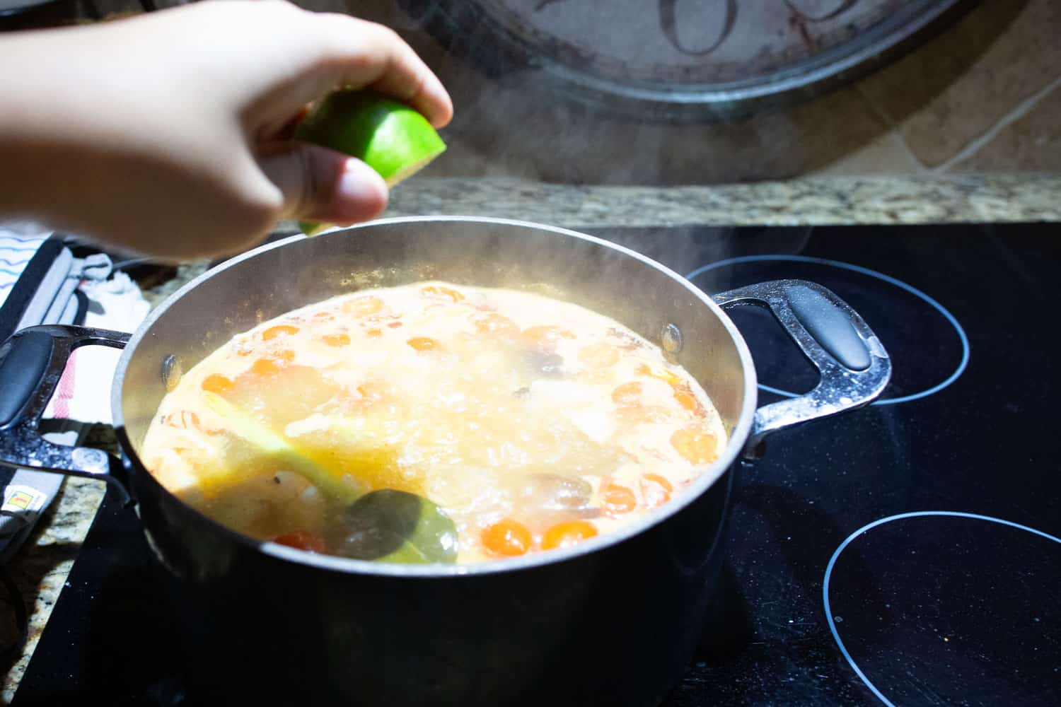A stainless steel pot on a stovetop with lemongrass, lime leaves, sliced galangal, and chilies simmering in clear broth