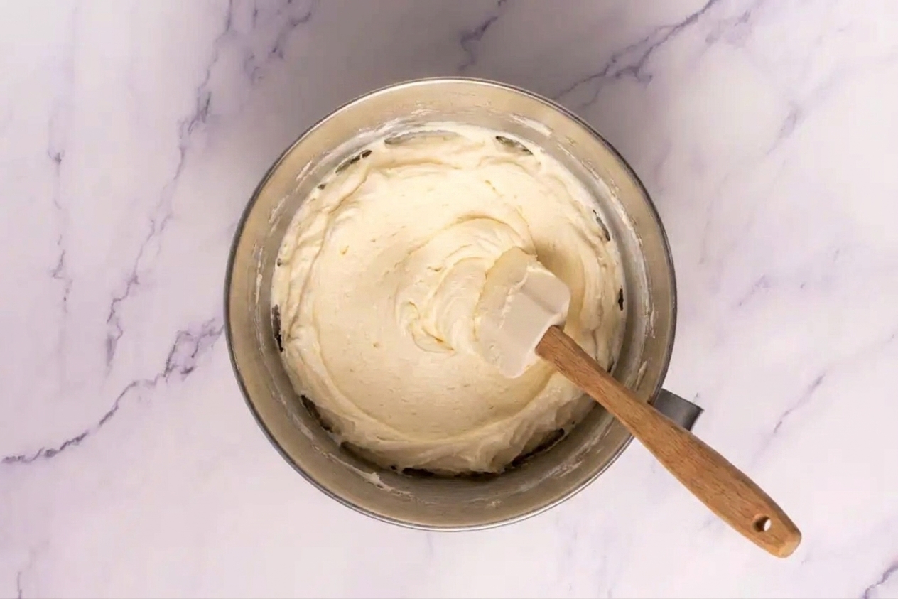 A stand mixer bowl filled with fluffy vanilla buttercream frosting with a spatula resting inside on a tidy kitchen counter