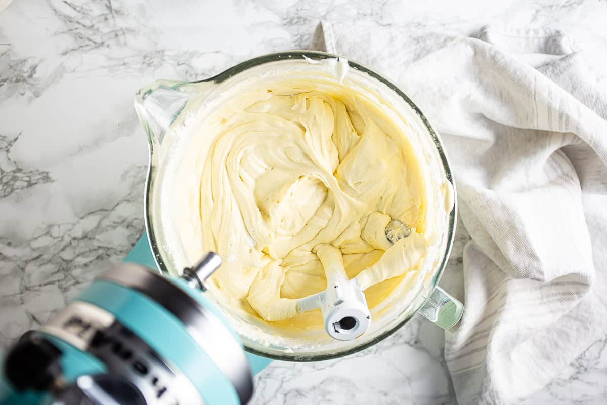A stand mixer bowl with cream cheese and butter being beaten smooth with a paddle attachment on a bright kitchen counter