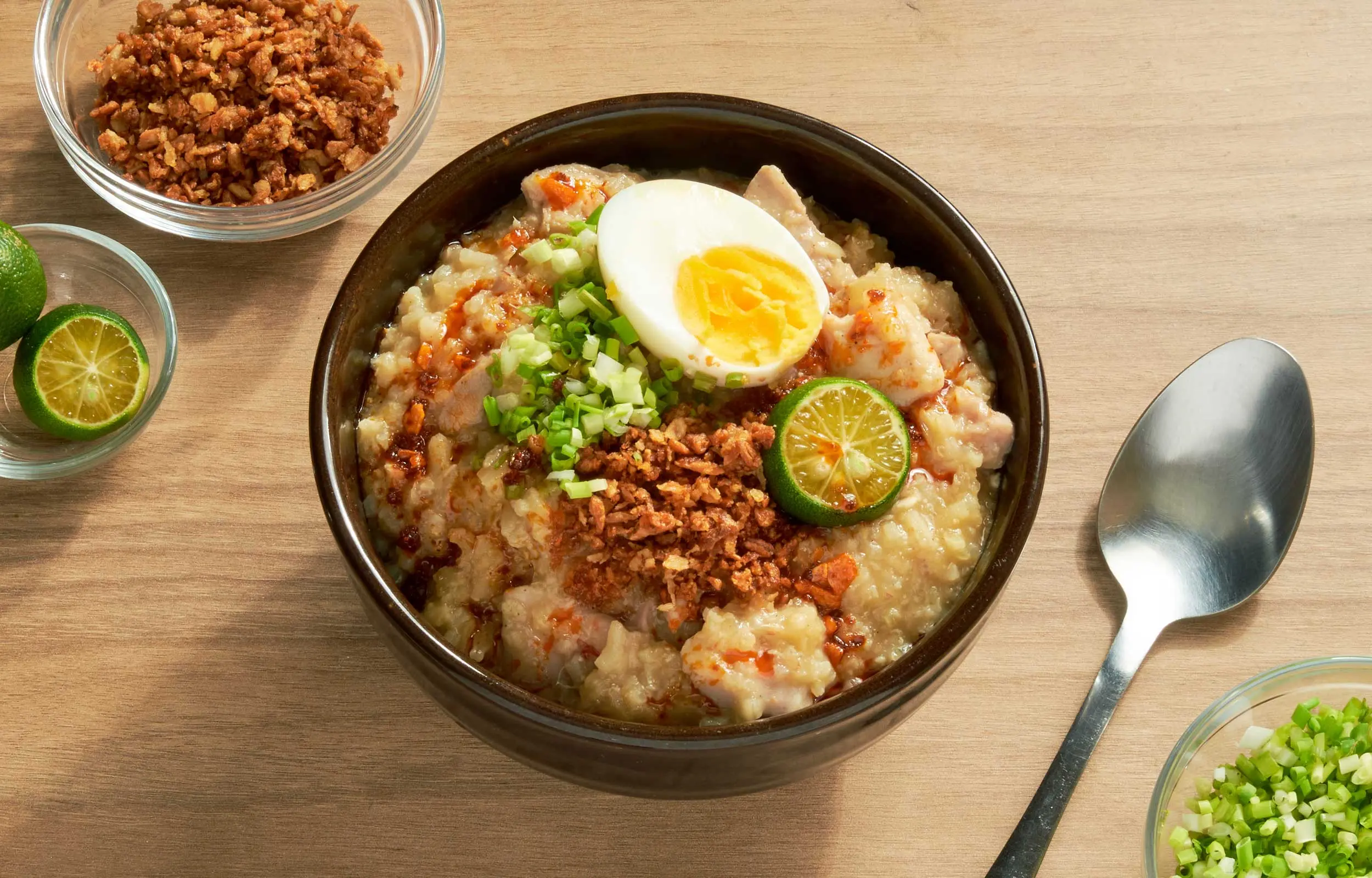A steaming bowl of Filipino arroz caldo chicken rice porridge topped with sliced scallions, toasted garlic, and a calamansi half on a neutral-toned kitchen table