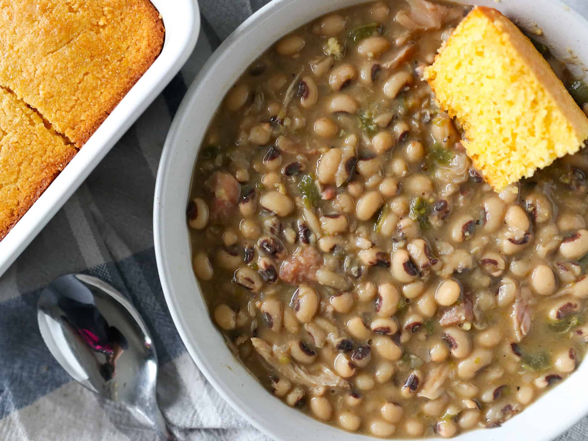 A steaming bowl of Southern black-eyed peas with visible ham pieces and chopped green onions, set on a rustic wooden table with cornbread nearby