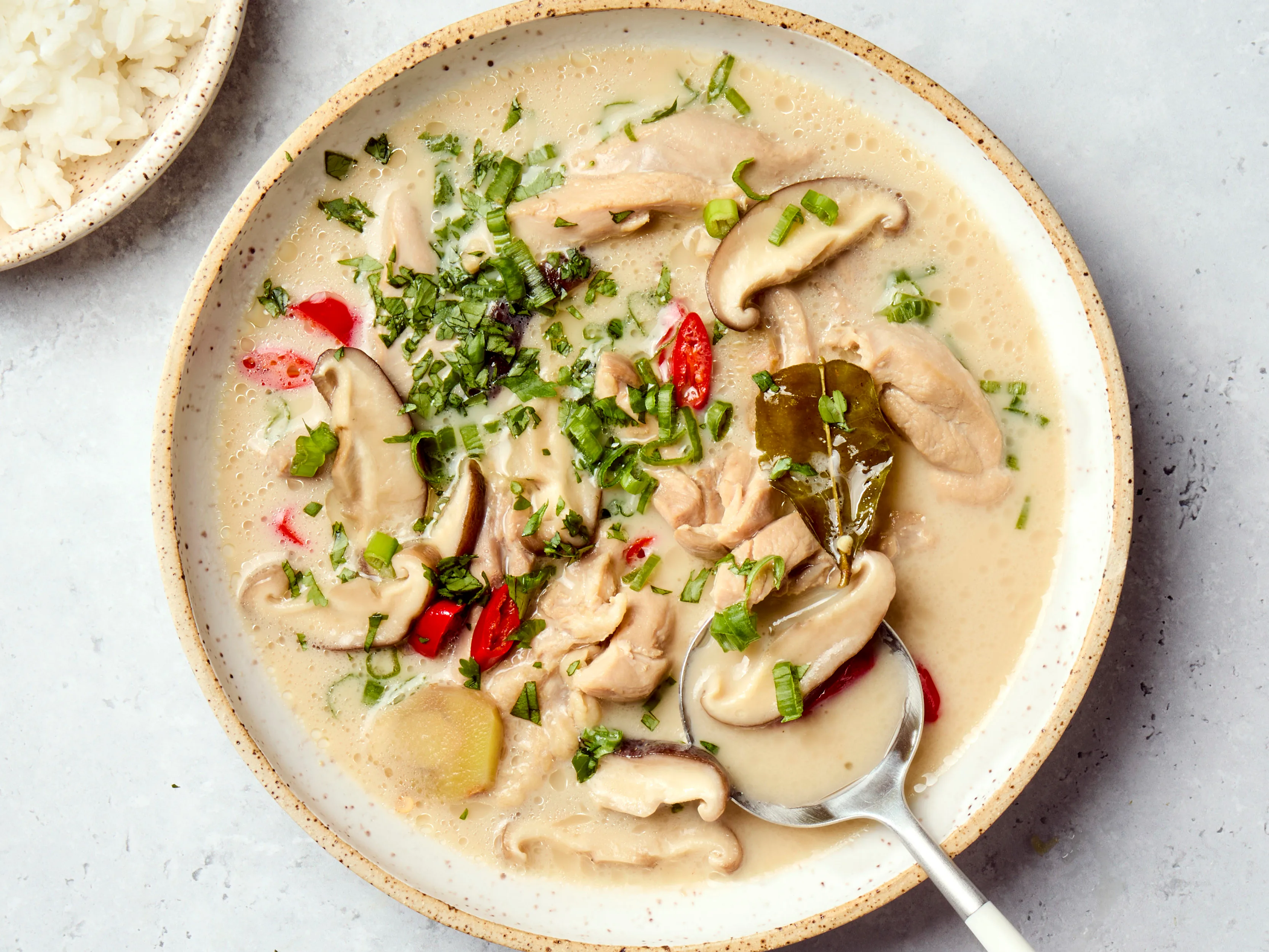 A steaming bowl of Thai coconut soup with chicken, mushrooms, cilantro, and sliced red chiles on a light kitchen table with lime wedges
