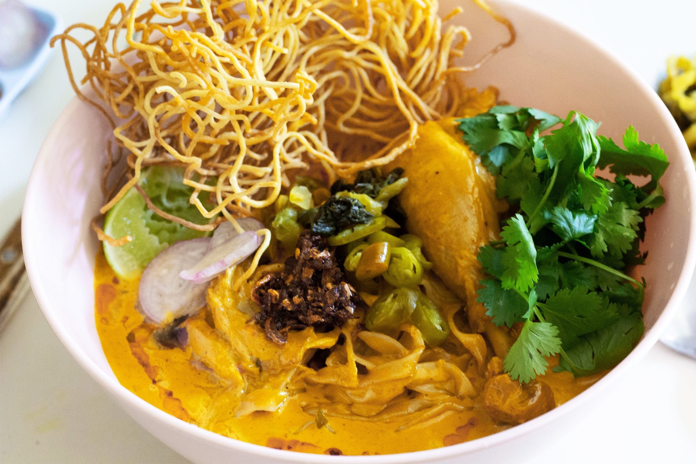A steaming bowl of Thai khao soi with golden coconut curry broth, tender chicken, soft egg noodles, crispy fried noodles on top, and lime wedges on a wooden table