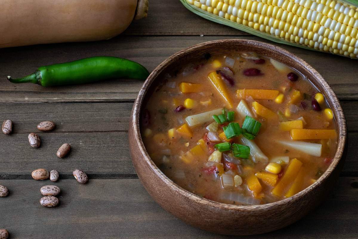 A steaming bowl of Three Sisters stew with chunks of squash, beans, and corn in a tomato-red broth, set on a wooden table with a simple spoon and napkin