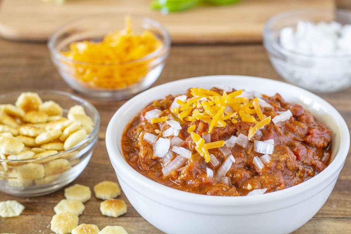 A steaming bowl of Wendy’s-style copycat chili topped with shredded cheddar and chopped onions on a wooden kitchen table with a spoon beside it