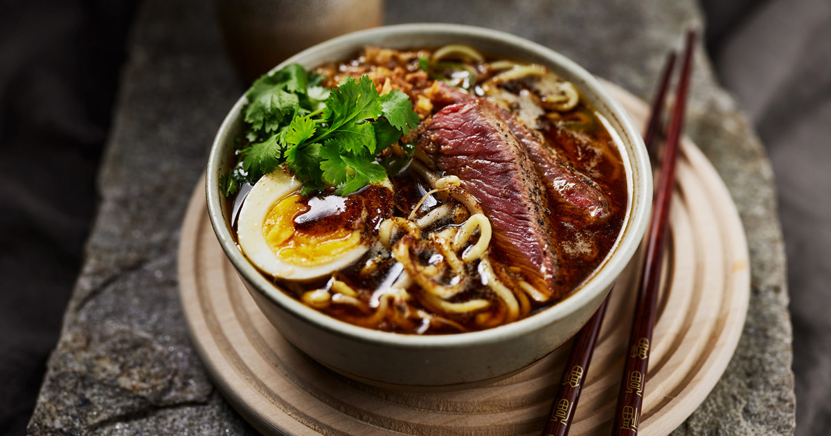 A steaming bowl of beef ramen with glossy noodles, sliced seared beef, scallions, and a soft-boiled egg on a wooden table with chopsticks nearby