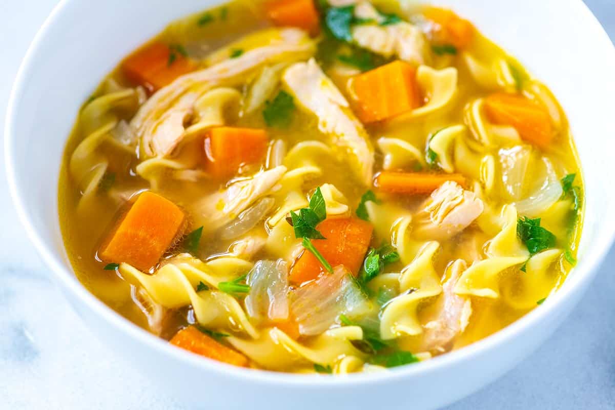 A steaming bowl of chicken and egg noodles in a golden broth with carrots, celery, parsley, and black pepper on a wooden table with a spoon beside it, warm natural kitchen lighting