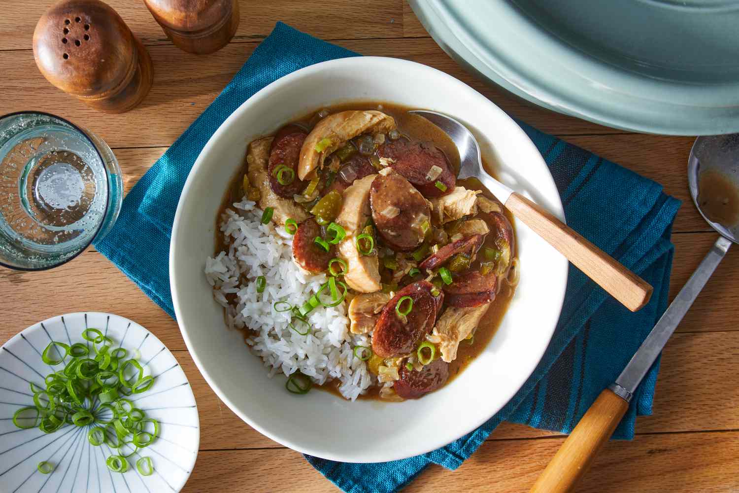 A steaming bowl of chicken and sausage gumbo with a dark, glossy broth, sliced sausage rounds, tender chicken pieces, and chopped parsley served next to white rice on a rustic table