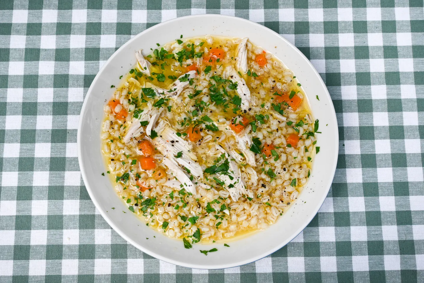 A steaming bowl of chicken barley soup with shredded chicken, carrots, celery, and pearled barley in a light golden broth on a wooden table with a spoon beside it