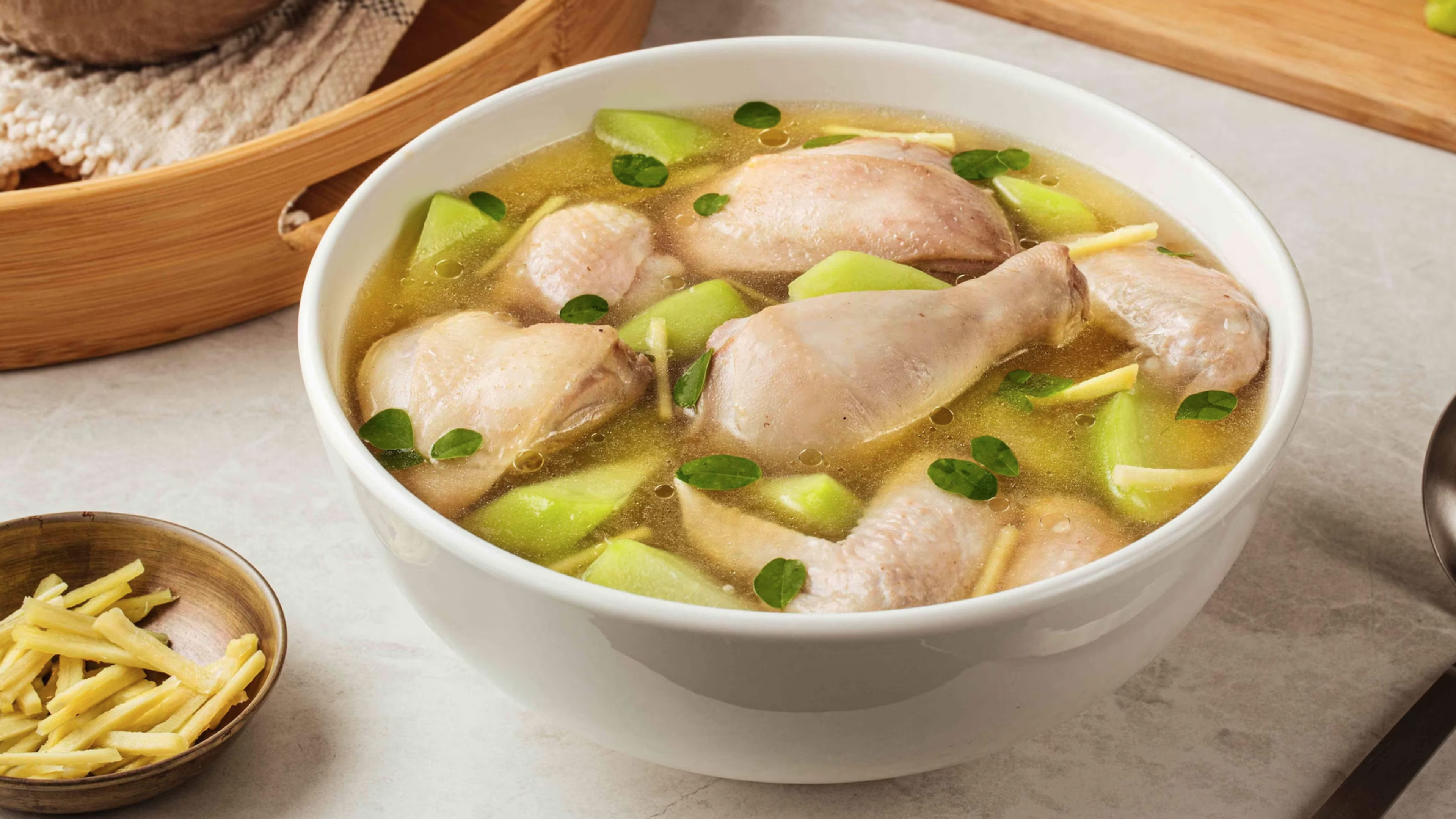 A steaming bowl of chicken tinola with clear ginger broth, chicken pieces, chayote slices, and leafy greens on a simple wooden table with soft natural light