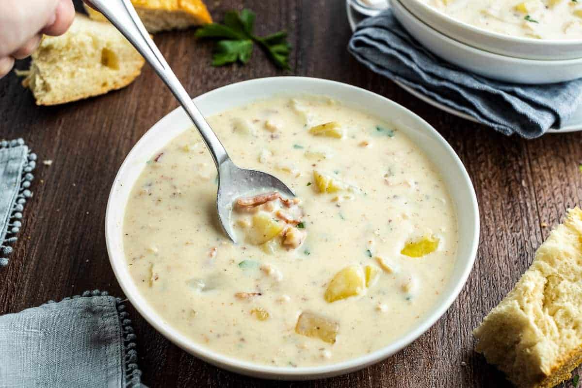 A steaming bowl of creamy New England clam chowder topped with crisp bacon and chopped parsley on a wooden table with oyster crackers nearby