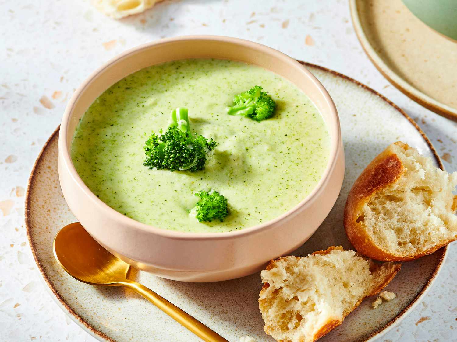 A steaming bowl of creamy broccoli soup topped with cracked black pepper and a small pile of shredded cheddar on a wooden table with a spoon beside it